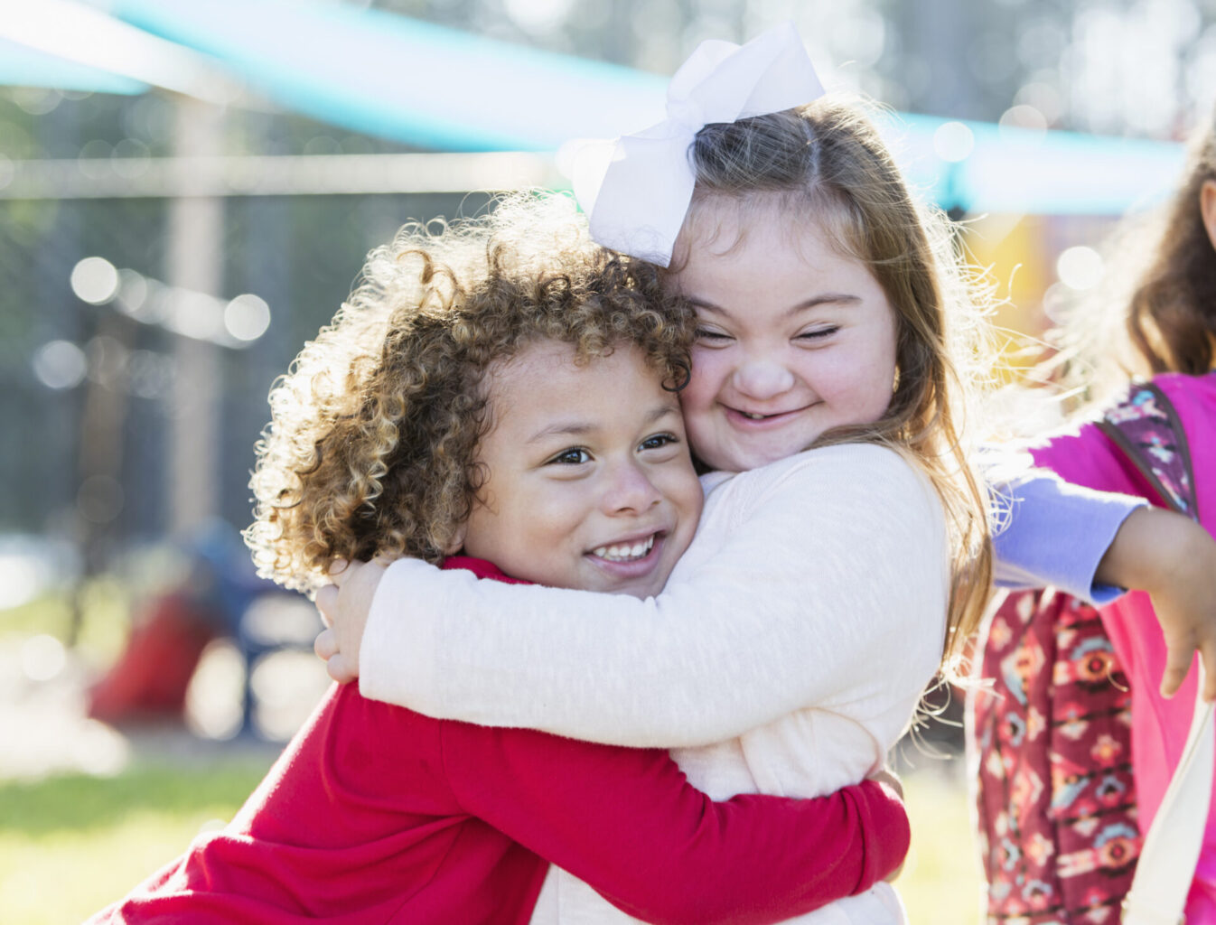 A pretty little girl with down syndrome and her cute 5 year old friend with curly hair, hugging each other and smiling.  The boy is mixed race African-American and Caucasian.